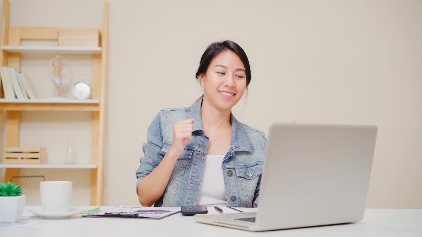 Woman sitting in front of a laptop, smiling with her hand closed and slightly raised, indicating a sense of achievement and joy