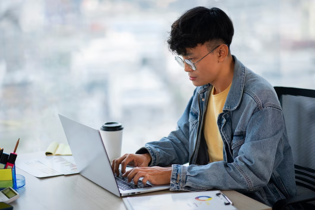 Man wearing glasses working on laptop at a table with a cup of coffee and documents nearby