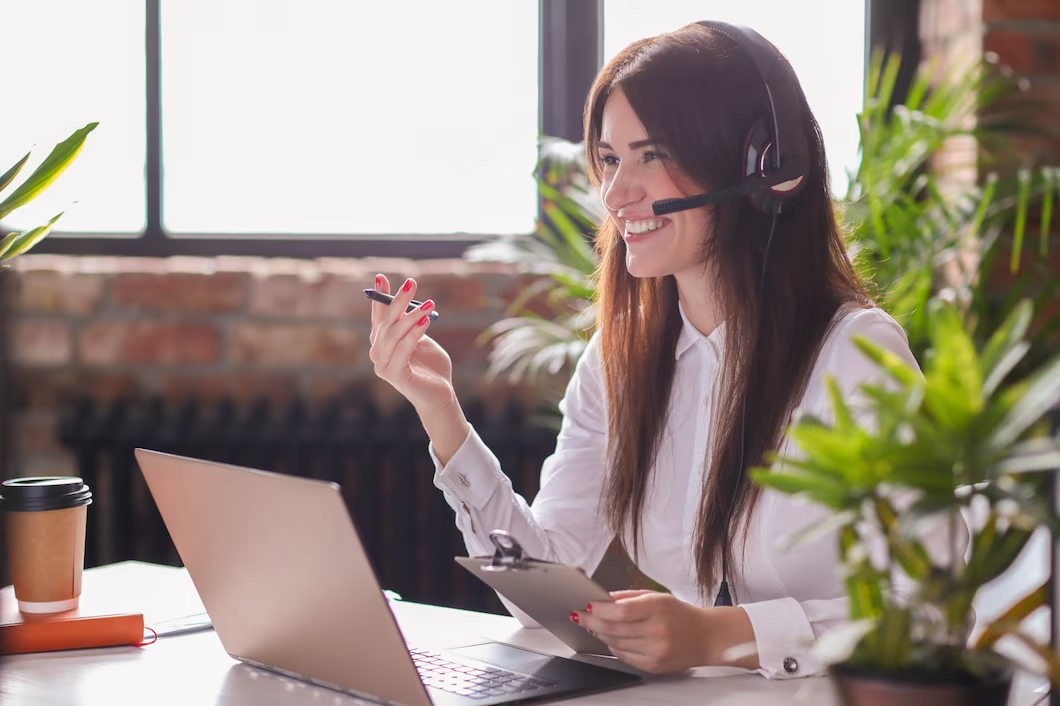 A smiling woman wearing a headset, holding a pen, and sitting in front of a laptop.